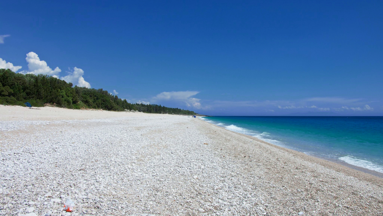 Dhërmi Beach Albania — beach clubs on the pebble shore with mountain backdrop and deep blue Ionian water